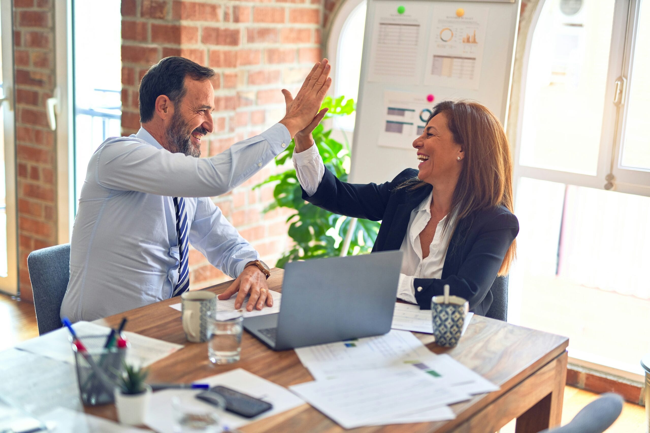 krakenimages-376KN_ISplE-unsplash Two business professionals celebrating success with a high-five at a modern office desk, surrounded by laptops, documents, and natural lighting